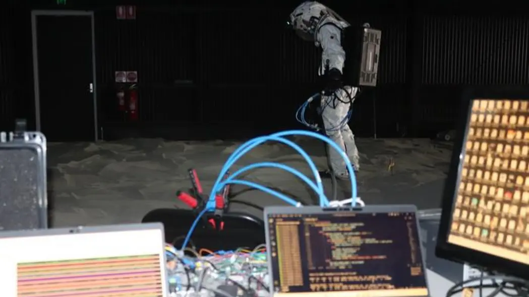 فردی که لباس سفیدی پوشیده و پشت میزی ایستاده است که سیم‌ها و رایانه‌های مختلفی روی آن قرار دارند