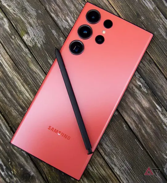 عکسی از یک Galaxy S23 Ultra قرمز که رو به پایین روی سطح چوبی قرار دارد و قلم S Pen روی آن قرار گرفته است