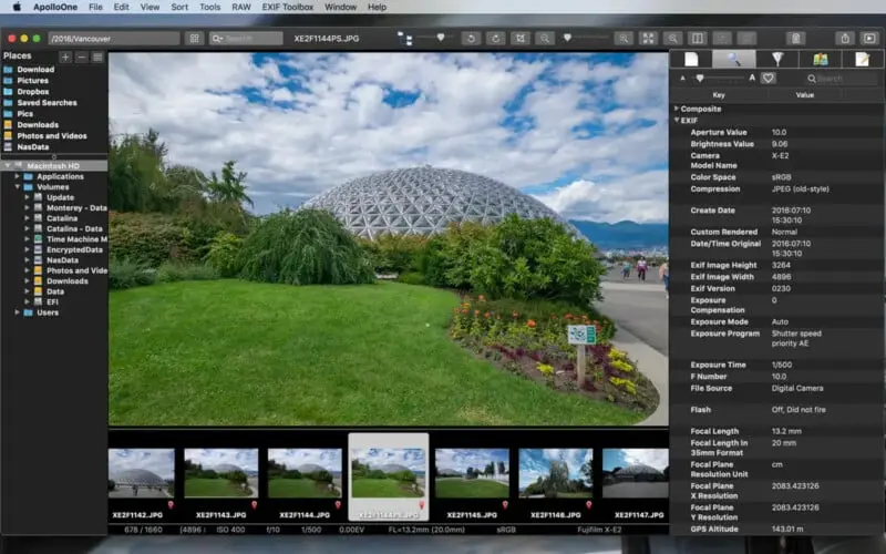 یک گلخانهٔ بزرگ با سقف شیشهای پشت چمن سبز و بسترهای گل رنگارنگ زیر آسمان نیمهابری قرار دارد؛ افراد در مسیر نزدیک قدم میزنند.