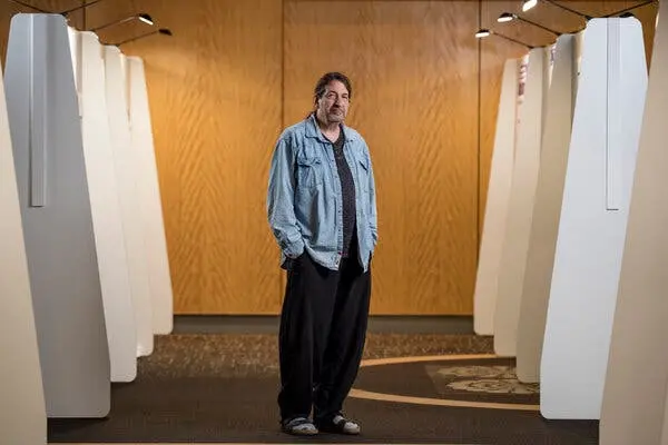 پرترهای از ریک استیونز که دستهایش را در جیبهایش دارد و در راهرویی با دیوارهای تقسیمکننده در هر دو طرف ایستاده است.