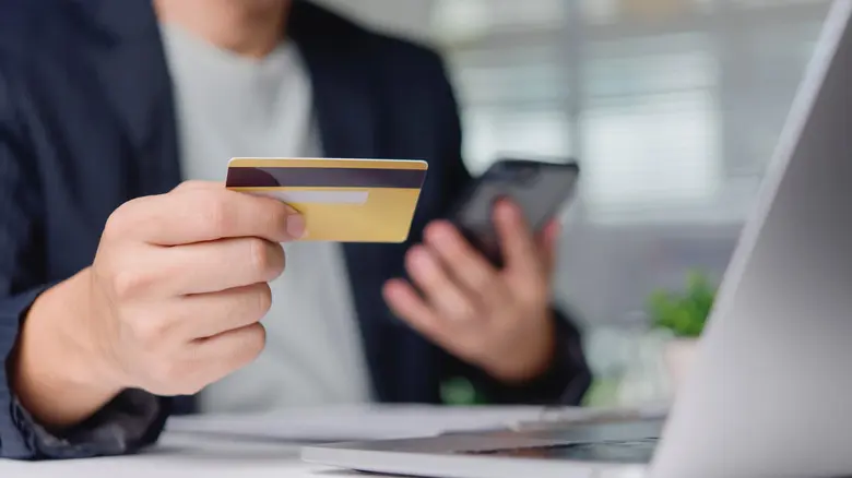 مشتری با کارت اعتباری و گوشی هوشمند خرید آنلاین را روی میز کار، کنار لپ‌تاپ تأیید می‌کند.