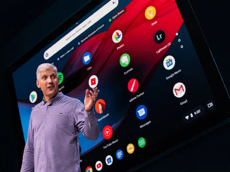 فردی با پیراهن بلندآستین بنفش که به صفحه‌نمایش بزرگ پشت سر خود اشاره می‌کند