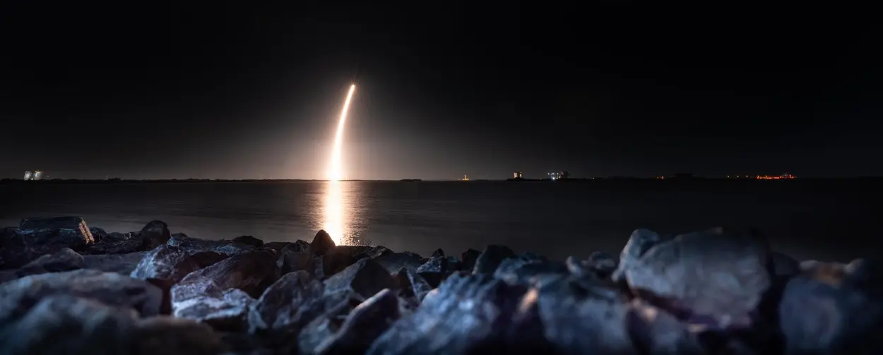 تصویری از آسمان شب با یک رد طلایی که از آب منعکس می‌شود، ناشی از پرتاب موشک SpaceX Falcon 9 حامل فرودگر Blue Ghost Mission One شرکت Firefly Aerospace که پس از پرتاب در پایگاه پرتاب ۳۹A مرکز فضایی کندی ناسا، فلوریدا، در روز چهارشنبه ۱۵ ژانویه ۲۰۲۵ به سمت بالا پر می‌کشد.
