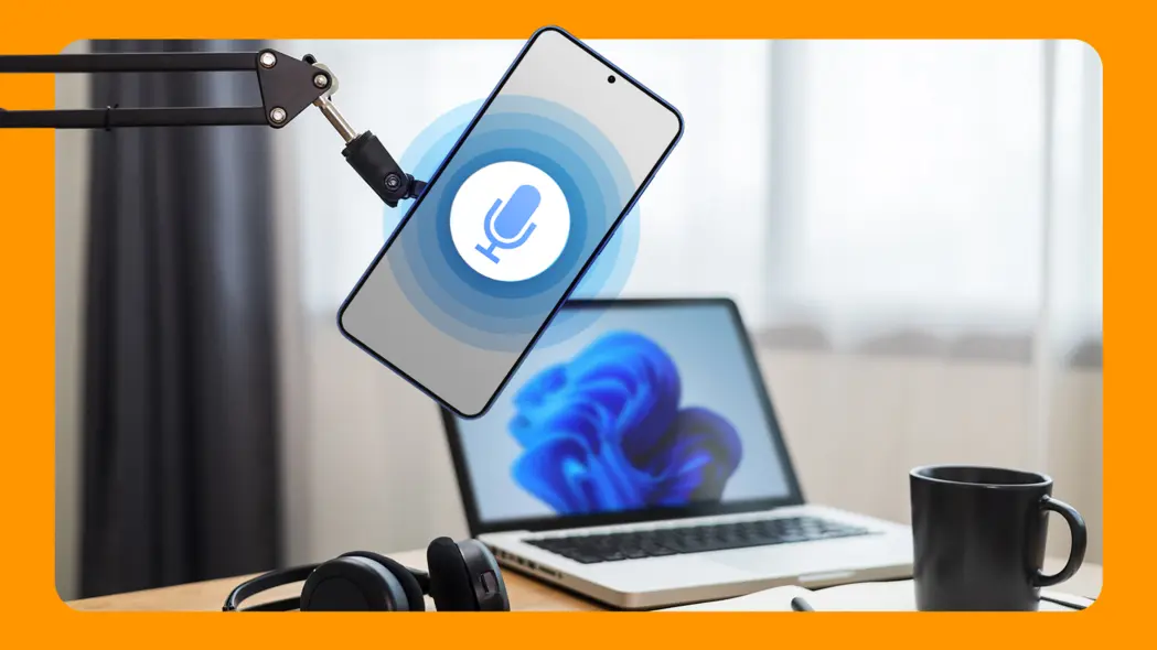 گوشی هوشمند با نماد میکروفن روی بازوی میکروفن، همراه با لپتاپ، هدفون و فنجان قهوه روی میز در پسزمینه.