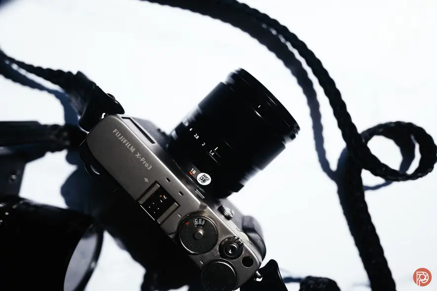 دوربین فوجیفیلم سری X با لنز و بند سیاه بر روی سطح سفید، از بالا عکاسی شده.