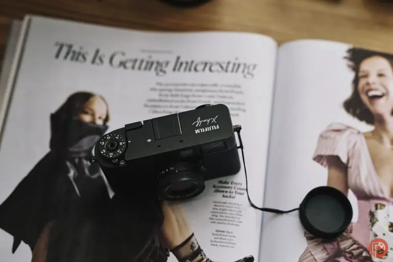 دوربین فوجیفیلم سری X بر روی یک مجله باز که حاوی عکسها و متن است، با درپوش لنز متصل شده، قرار دارد.