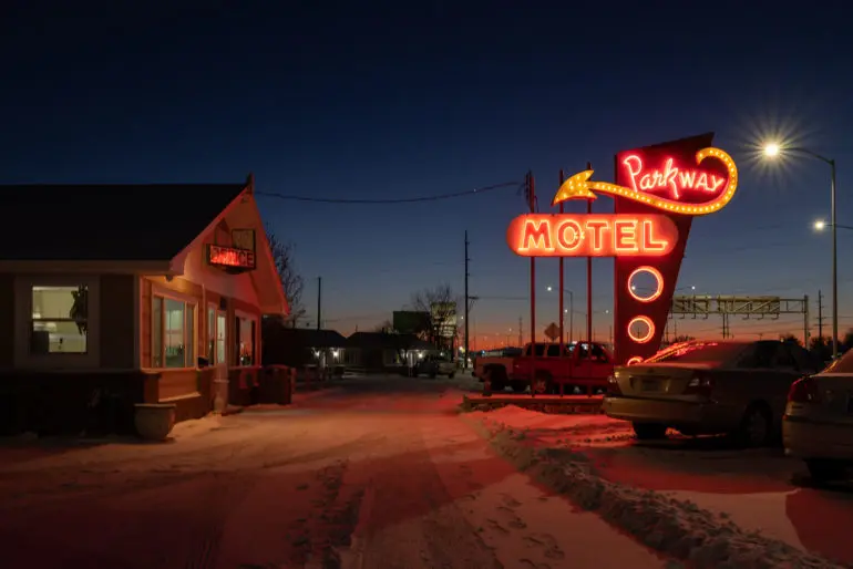 علامت نئونی Motel Parkway در زمان غروب در کنار خودروها و یک ساختمان کوچک در جادهٔ پوشیده از برف می‌درخشد.