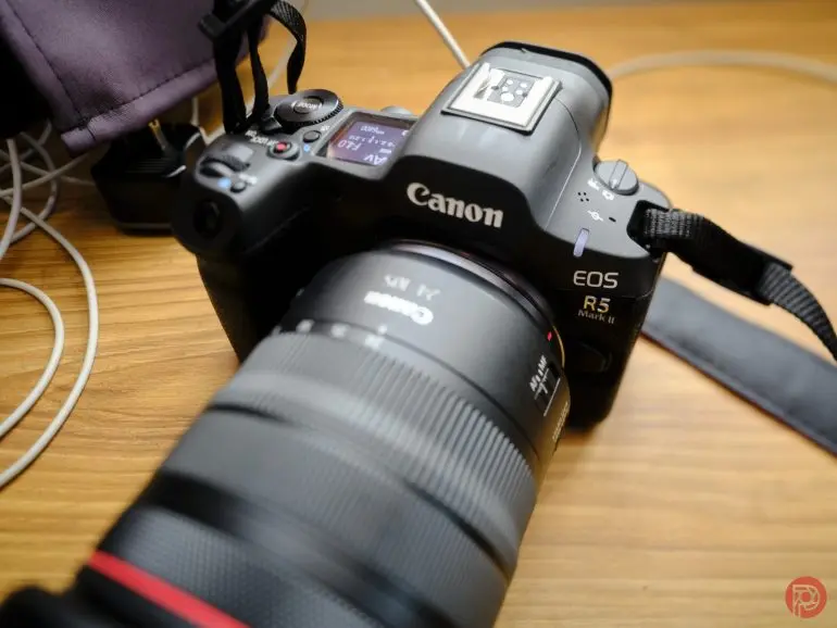 دوربین Canon EOS R5 با لنز بزرگ که روی سطح چوبی کنار چند سیم و یک کیف قرار دارد.