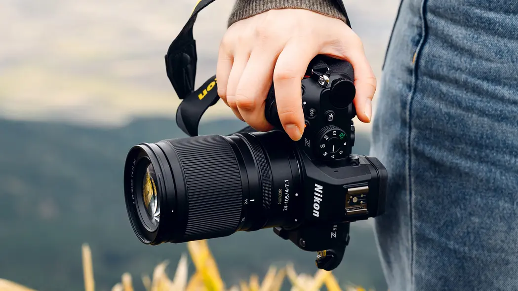 لنز Nikon Z 24‑105mm بر روی دوربین Nikon، در دست فردی با شلوار جین آبی و پس‌زمینهٔ روستایی